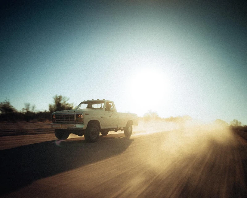 An older white pickup truck is driving on a dirt road with dust rising in the sunlight under a clear blue sky in a rural environment.