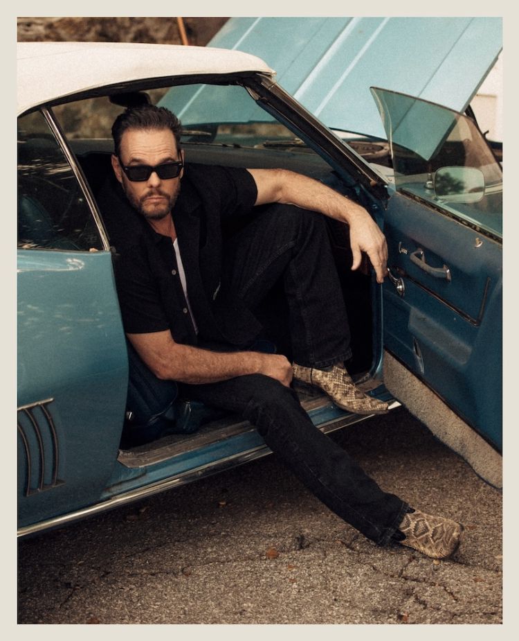Adult male poses in classic car with open hood wearing black shirt, dark jeans, sunglasses, and snakeskin boots. Outdoors setting.
