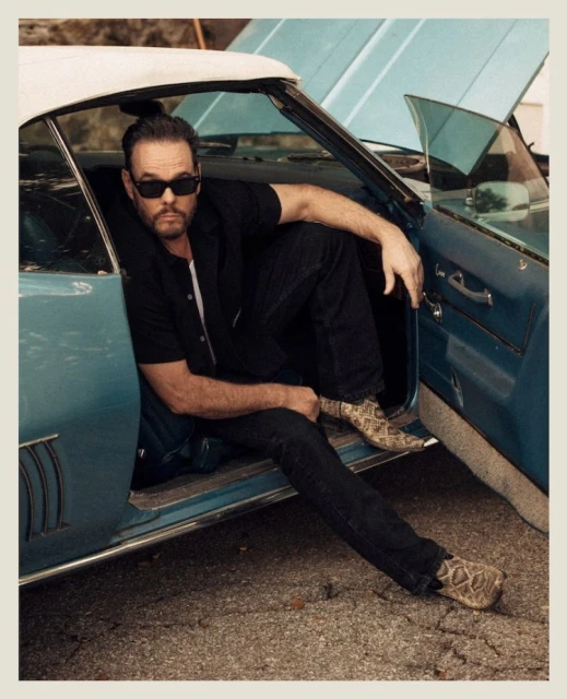 Adult male poses in classic car with open hood wearing black shirt, dark jeans, sunglasses, and snakeskin boots. Outdoors setting.