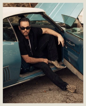 Adult male poses in classic car with open hood wearing black shirt, dark jeans, sunglasses, and snakeskin boots. Outdoors setting.