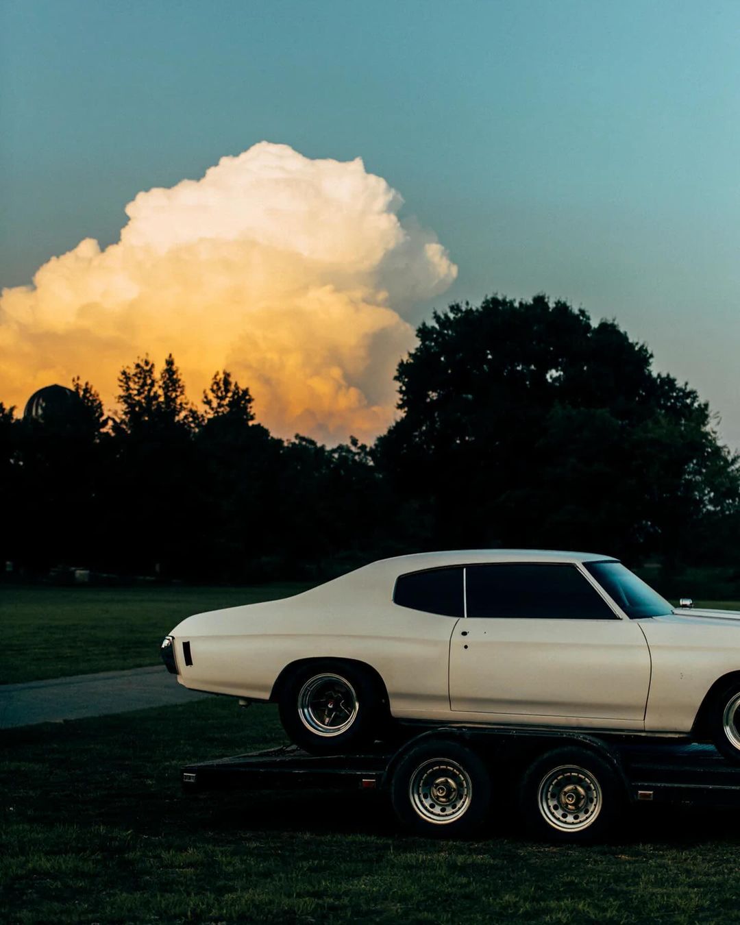 Ivory 1970s Chevelle coupe car on a trailer with a cloudscape background, parked on a grassy field at dusk, casting a dark mood.