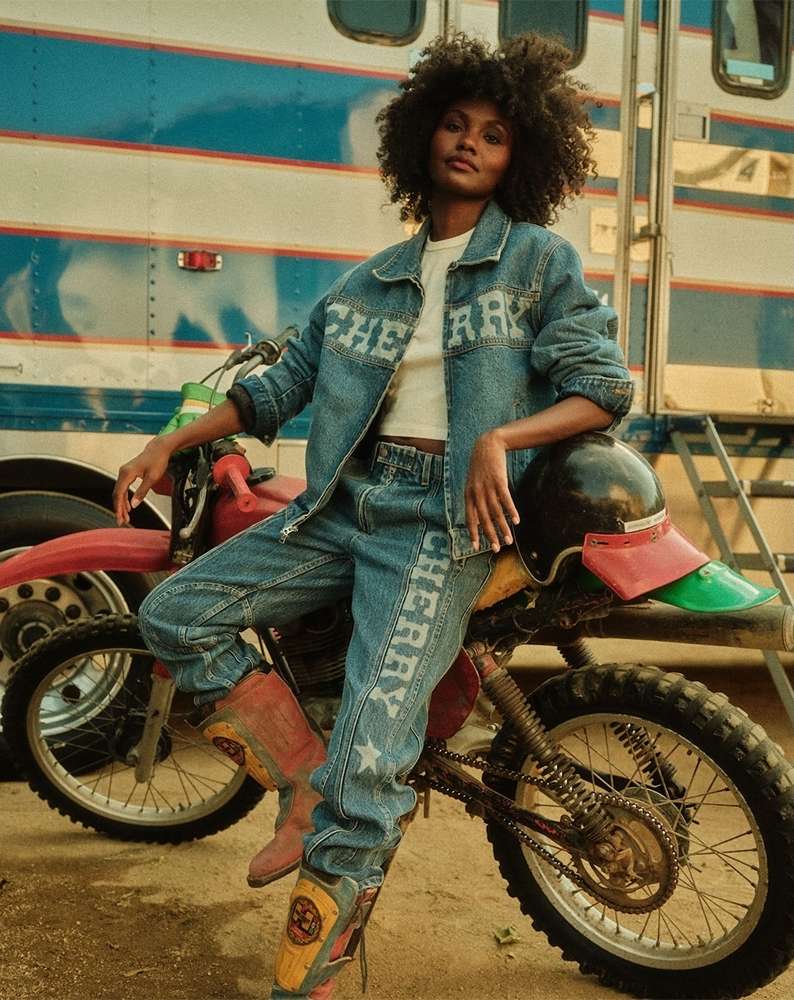 A Black woman in a denim jacket and pants sits on a motorcycle in front of a trailer, showcasing fashion and outdoor lifestyle.