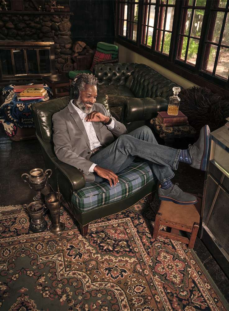 A mature Black man relaxes in an armchair in a sunlit room wearing headphones, a blazer, and jeans in an upscale, casual lifestyle image.