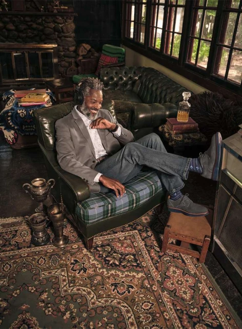 A mature Black man relaxes in an armchair in a sunlit room wearing headphones, a blazer, and jeans in an upscale, casual lifestyle image.