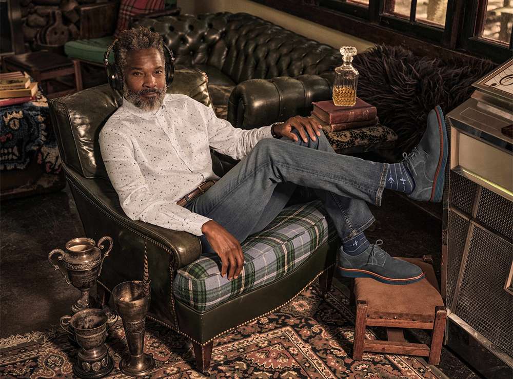 Man in armchair wearing headphones, dressed in button-down shirt and jeans, with feet on stool, in a stylish, vintage-inspired interior.
