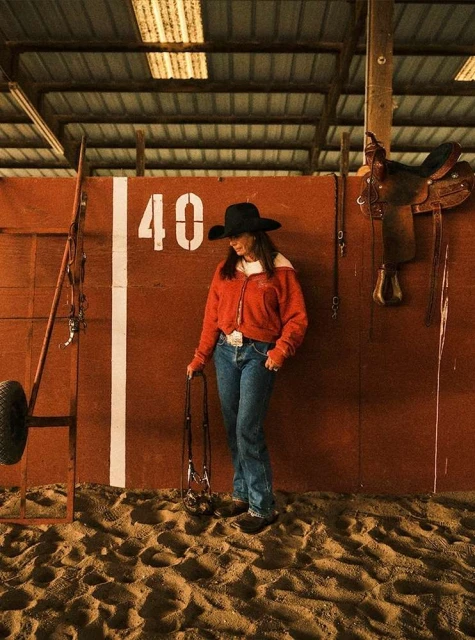 Adult female in cowboy hat stands in an indoor arena with sandy ground. Saddle, horse tack and horse training equipment in the background.