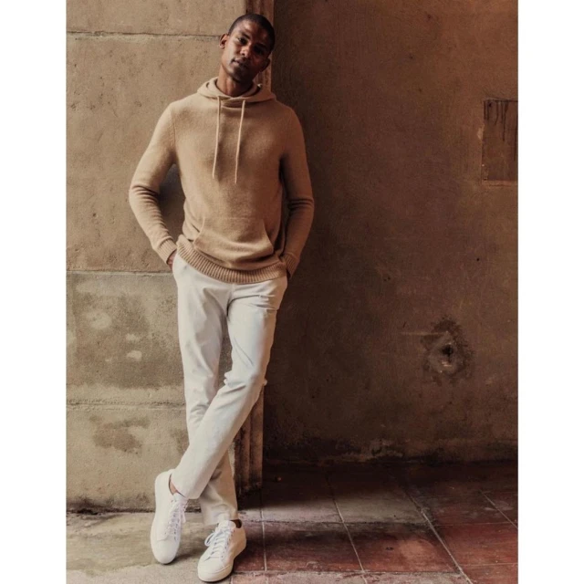 Man wearing a tan hooded sweatshirt and white pants leans against a wall in a medium shot. He is wearing white sneakers.