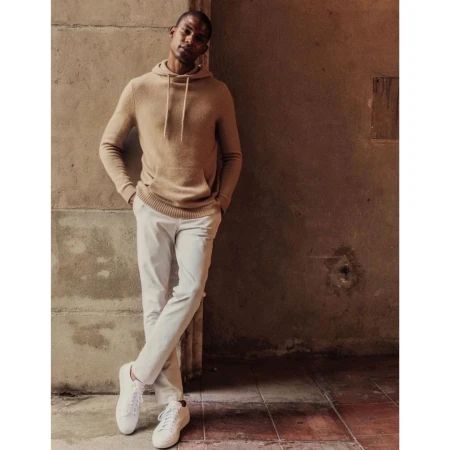 Man wearing a tan hooded sweatshirt and white pants leans against a wall in a medium shot. He is wearing white sneakers.