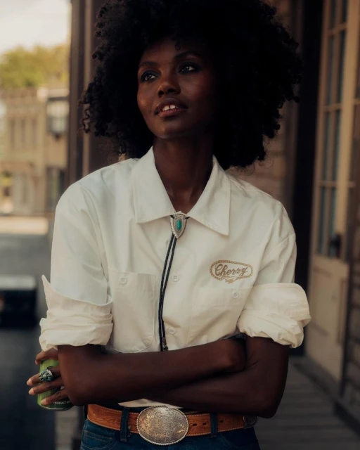 Portrait of a black woman with afro posing outdoors with folded arms, wearing a white shirt, bolo tie, belt buckle and holding a can.