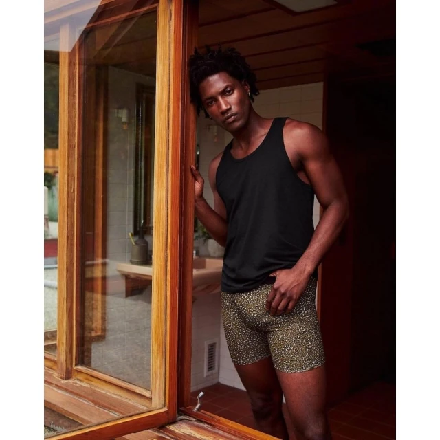 An African American male stands indoors near a wooden frame in a black tank top and patterned shorts, looking at the camera.