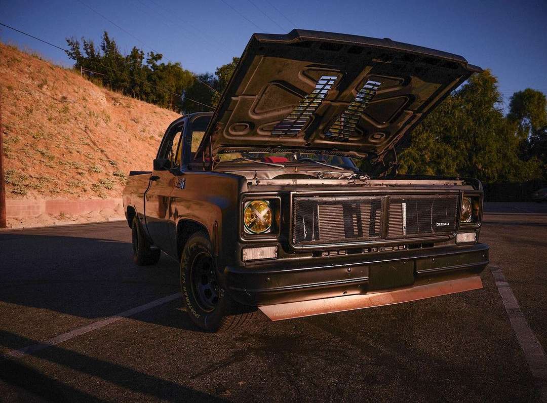 Front-angle view of a customized black pickup truck with its hood open, parked on asphalt against a backdrop of a grassy hill.