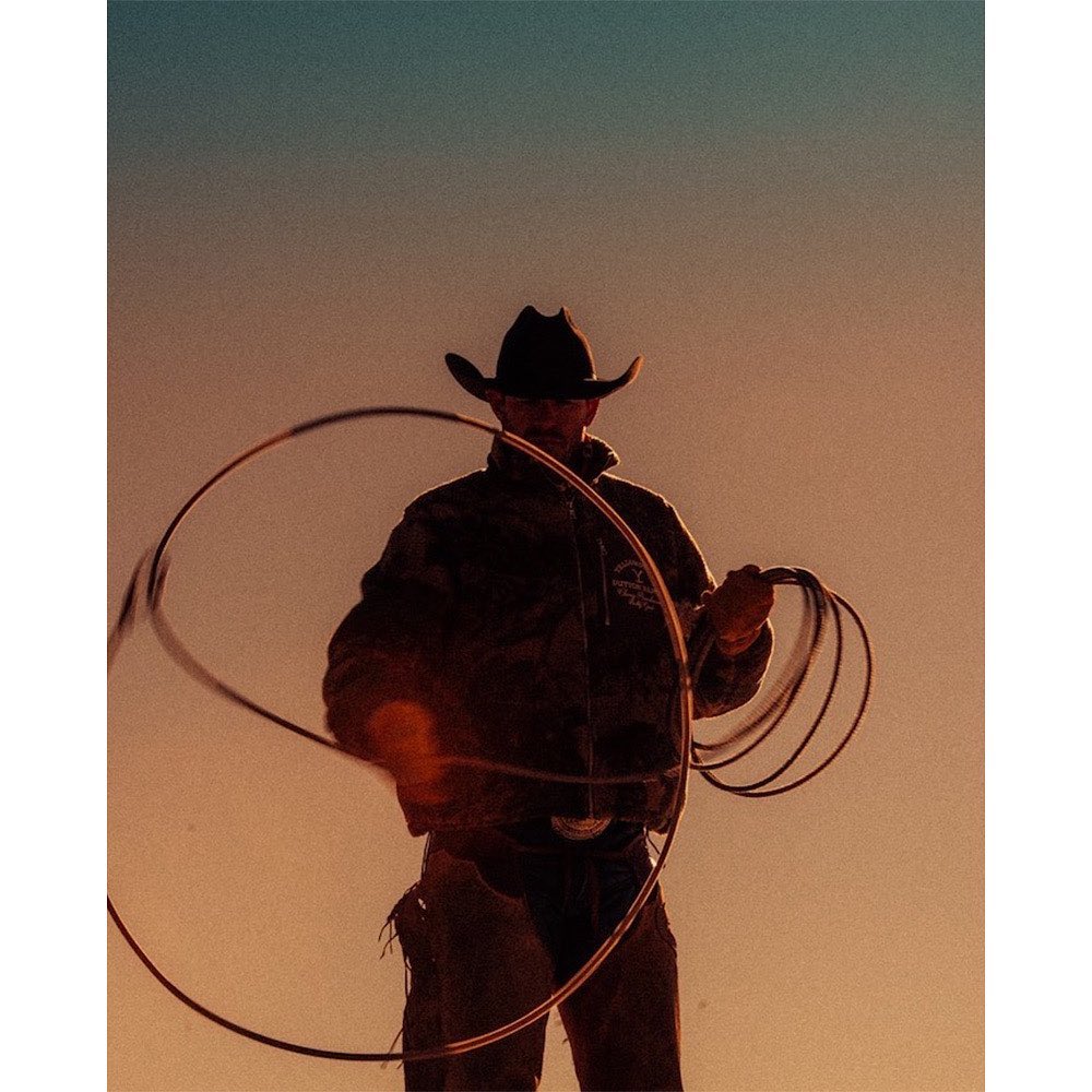 Silhouette of a cowboy in a hat twirling a lasso against a colorful sky at dusk, showcasing the Western lifestyle and rural setting.