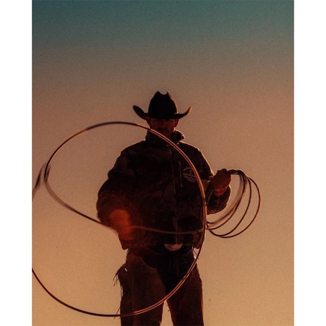 Silhouette of a cowboy in a hat twirling a lasso against a colorful sky at dusk, showcasing the Western lifestyle and rural setting.