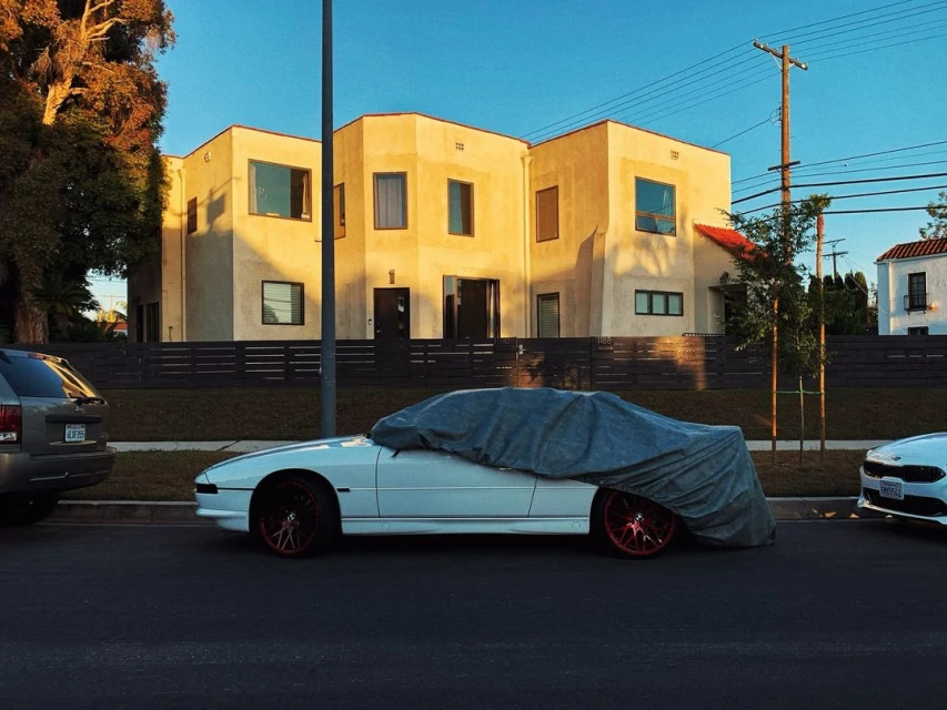 A white car is covered by a grey car cover on the side of a city street with a tan building behind it under a clear blue sky.