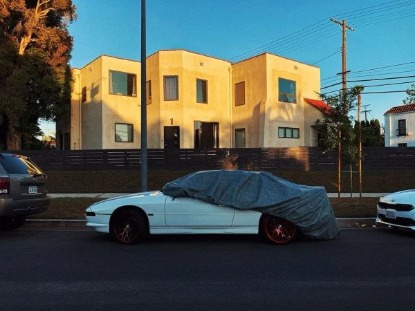 A white car is covered by a grey car cover on the side of a city street with a tan building behind it under a clear blue sky.