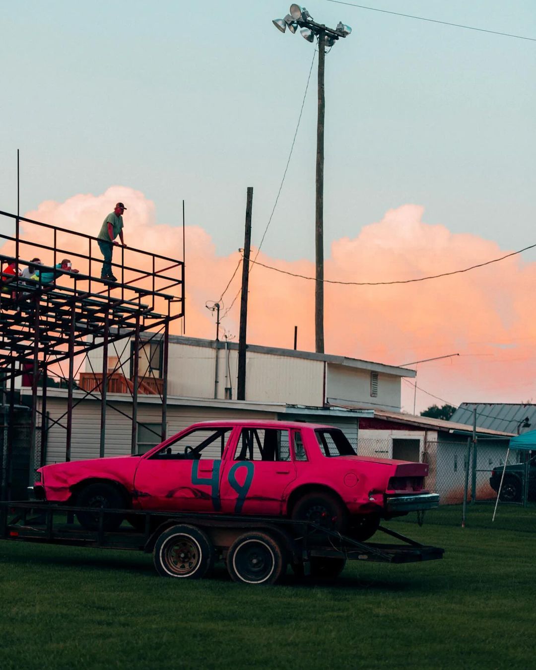 Bright pink demolition derby car with a large number 49 sits on a trailer; men stand on an elevated platform on a late afternoon.