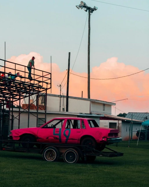 Bright pink demolition derby car with a large number 49 sits on a trailer; men stand on an elevated platform on a late afternoon.