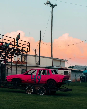 Bright pink demolition derby car with a large number 49 sits on a trailer; men stand on an elevated platform on a late afternoon.
