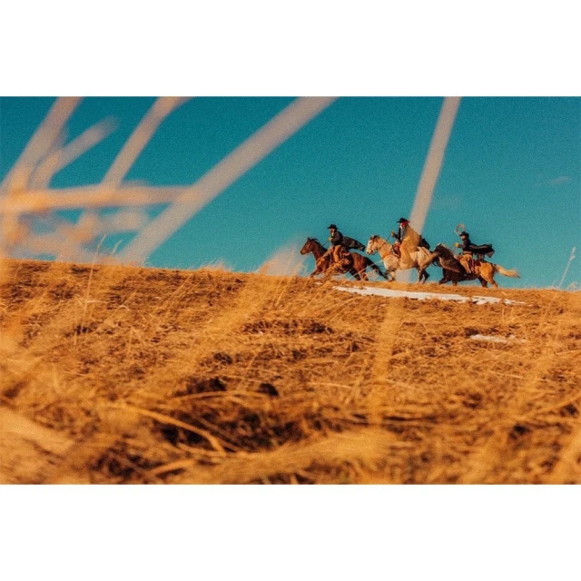 Four horseback riders gallop across a grassy hilltop against a clear blue sky, conveying a sense of speed, freedom, and outdoor adventure.