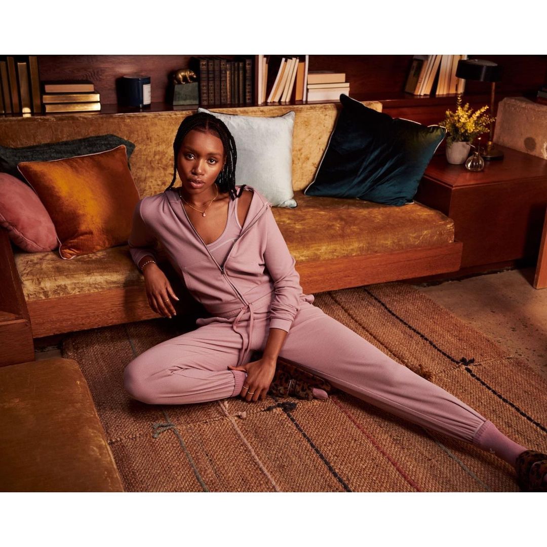An adult female with braided hair is sitting on a braided rug in a mauve-colored jumpsuit in front of a gold velvet couch and pillows.