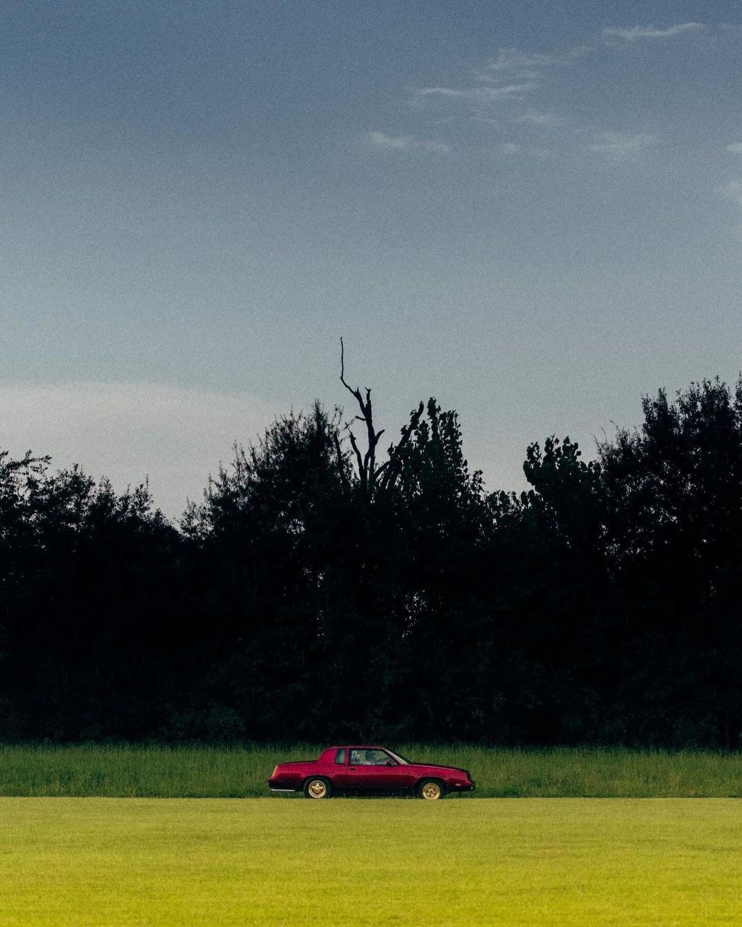 A maroon vintage car is parked in a field of green grass with a treeline in the background beneath a cloudy blue sky.