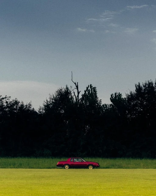 A maroon vintage car is parked in a field of green grass with a treeline in the background beneath a cloudy blue sky.