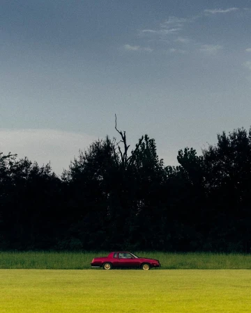 A maroon vintage car is parked in a field of green grass with a treeline in the background beneath a cloudy blue sky.
