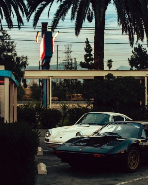 Two classic cars, a white Porsche and a black Ferrari, are parked in a lot with palm trees and a vintage sign in the background.