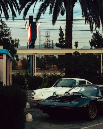 Two classic cars, a white Porsche and a black Ferrari, are parked in a lot with palm trees and a vintage sign in the background.