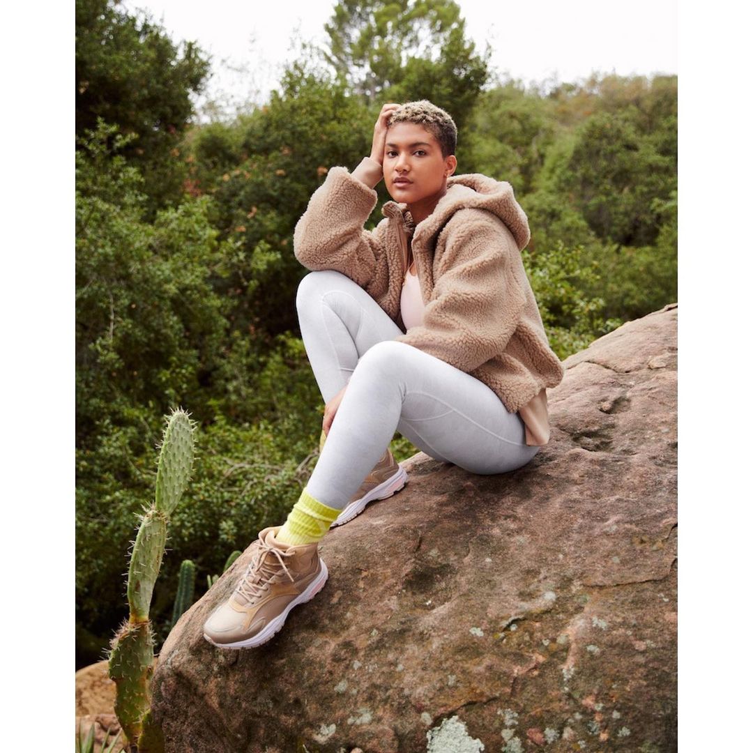 Woman in sherpa jacket and leggings sitting on rock in nature setting with trees in background, wearing hiking shoes and yellow socks.