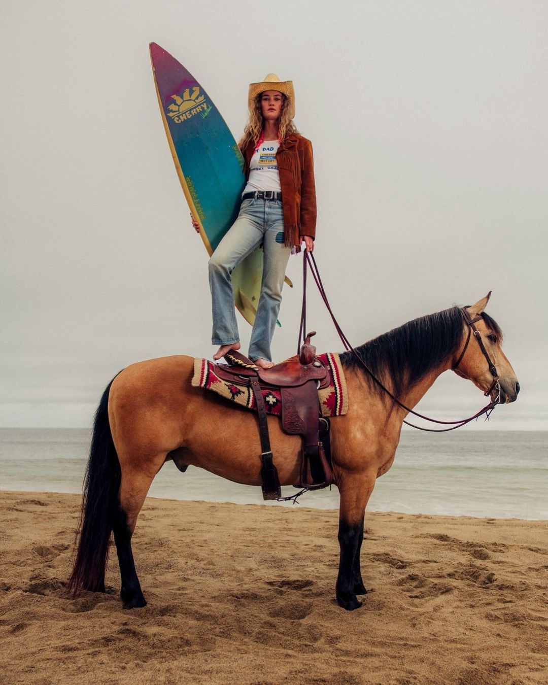 Woman wearing a cowboy hat standing on a horse on a sandy beach holding a colorful surfboard