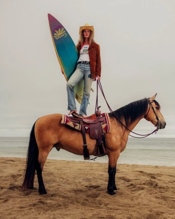 Woman wearing a cowboy hat standing on a horse on a sandy beach holding a colorful surfboard