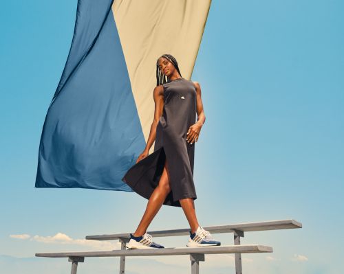 A woman in a black dress stands on a bench, showcasing athletic shoes against a blue sky with a billowing blue and tan backdrop.
