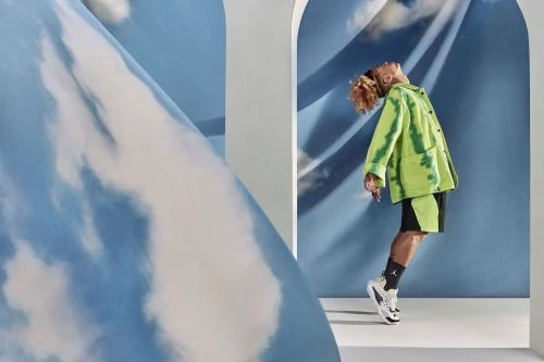 A person in a neon-green tie-dye jacket and shorts jumps in front of a blue cloud-patterned backdrop, framed by white arched doorways.