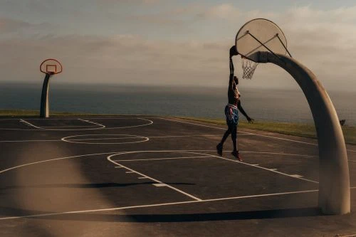 An athlete dunks a basketball outdoors, the sea visible in the background, creating a dynamic shot of skill and athleticism.