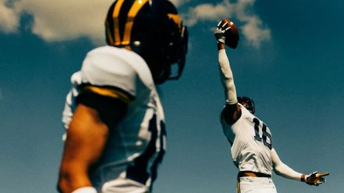 Two football players in uniform on a sunny day. One player with number 16 raises his arm to catch a football against a blue sky.