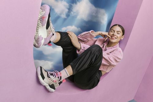 Adult woman wearing sneakers and a pink jacket smiles while reclining against a curved wall with a cloud-filled sky in the background.