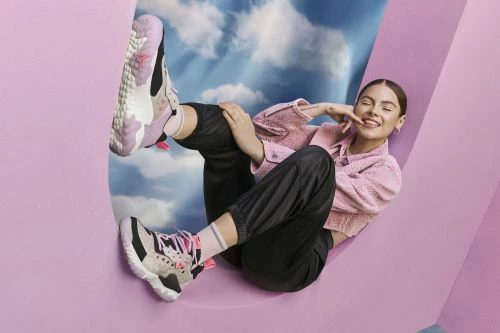 Adult woman wearing sneakers and a pink jacket smiles while reclining against a curved wall with a cloud-filled sky in the background.