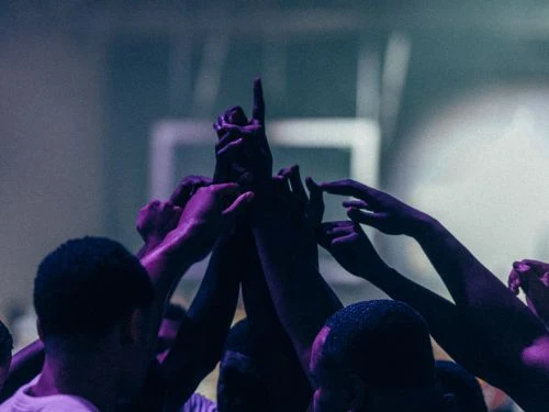 Group of people reaching upwards indoors, celebrating, teamwork, unity, indoor lighting with muted tones of blue, purple and gray.