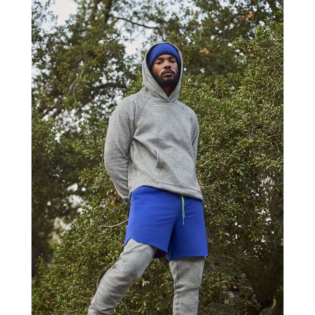 Man in grey hoodie and blue shorts stands outdoors against a backdrop of green foliage, showcasing casual athletic wear and natural setting.