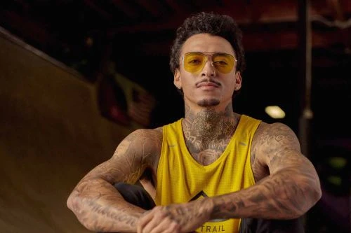 Man with tattoos in yellow tank top and sunglasses sits with arms crossed in an indoor setting, showcasing athletic fitness and style.