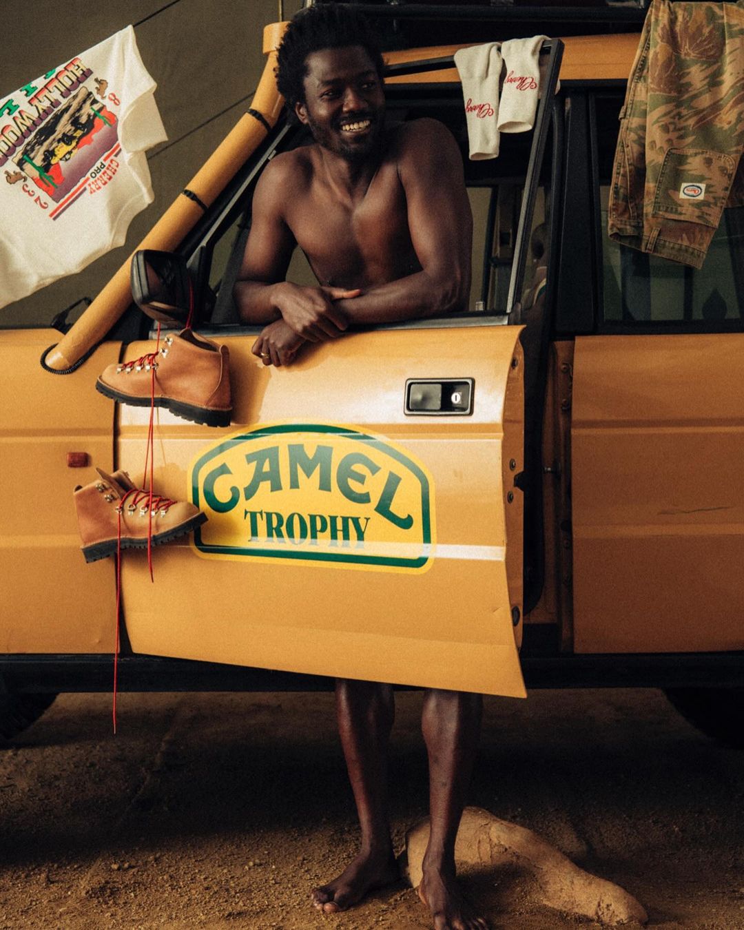 Man with tanned hiking boots leaning against a yellow SUV. Two shirts are hanging from the vehicle. Camo pants are also on display.