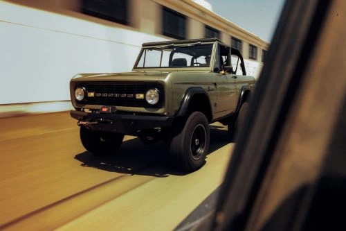Olive green Ford Bronco driving on a paved surface, captured with a shallow depth of field. Building is visible in the background.
