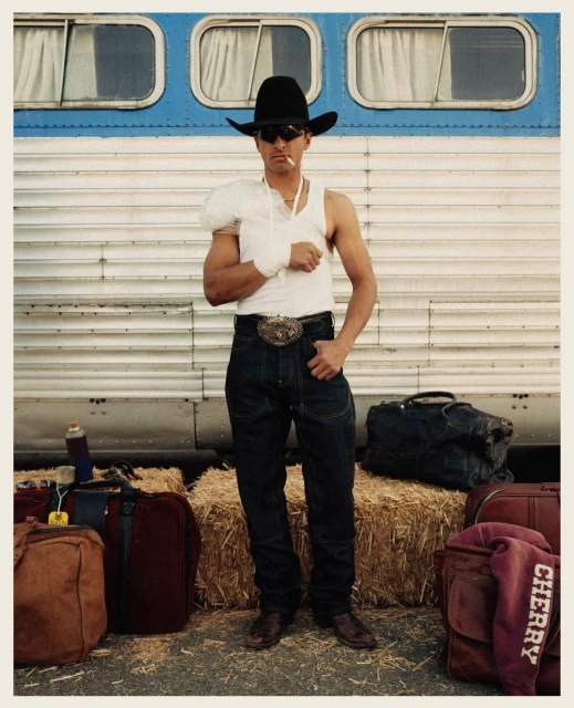 Cowboy with a cigarette in his mouth stands in front of a blue and silver bus with hay bales and luggage at his feet.