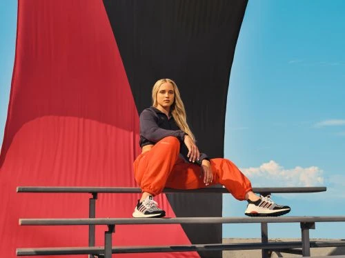 Athletic woman poses on railing in orange pants and black athletic top in an outdoor setting with a large red and black backdrop.