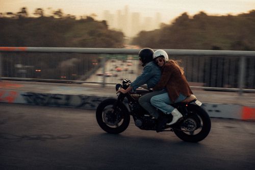 Two people on a black Honda motorcycle riding over a bridge with a city skyline in the background at sunset, outdoor travel lifestyle.