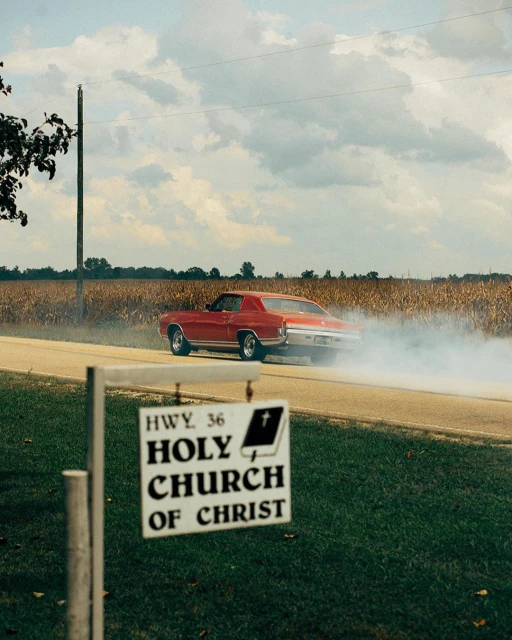 Classic red muscle car drifting on a rural road near a cornfield and church sign on a sunny day, creating a dynamic and nostalgic scene.