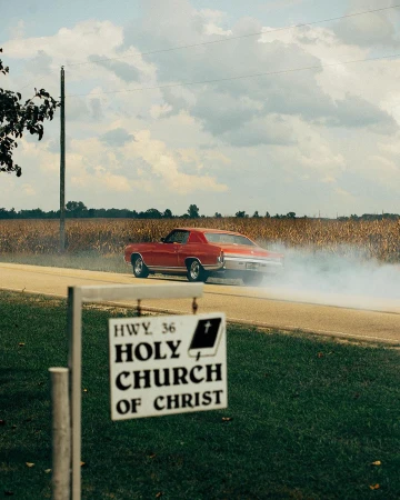 Classic red muscle car drifting on a rural road near a cornfield and church sign on a sunny day, creating a dynamic and nostalgic scene.