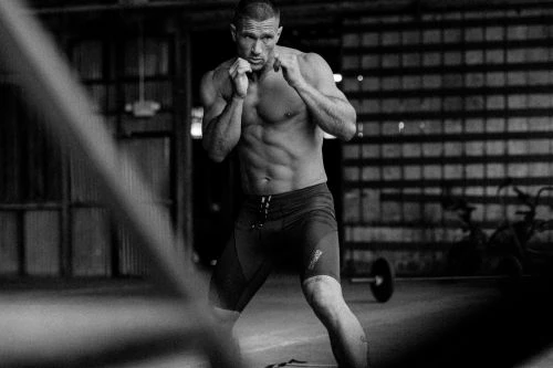 Black and white shot of a muscular adult male in athletic shorts posing with fists up in an indoor gym setting.