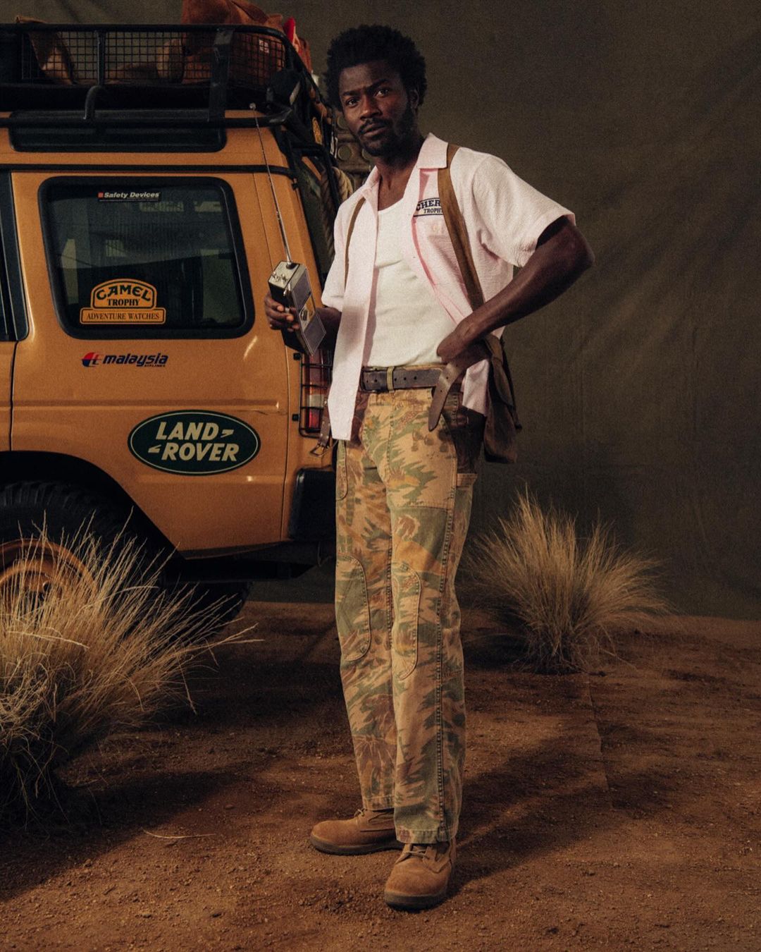 Man in retro attire poses with a vintage radio next to a classic Land Rover, creating a nostalgic and adventurous scene with desert backdrop.